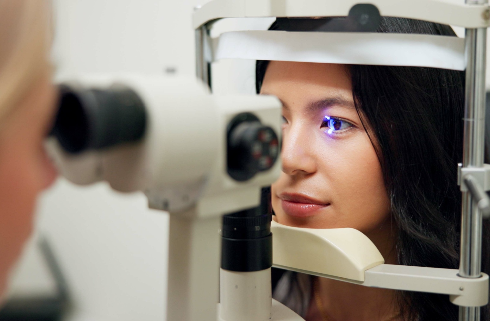 A close-up image of a young woman sitting in a chair, facing an optometrist during her eye exam. A bright light is being shone into her eye.