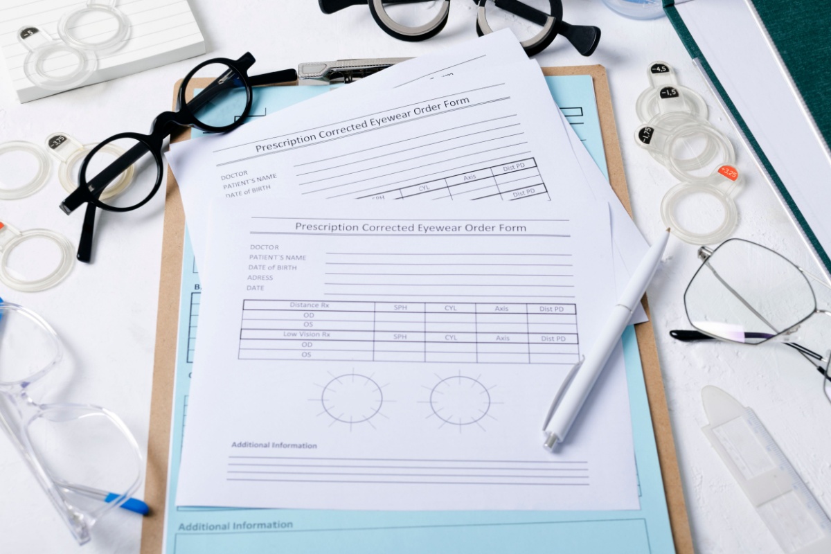 Prescription eyewear order forms on a clipboard surrounded by eyeglasses, trial lenses, and optical tools used for contact lens fittings.