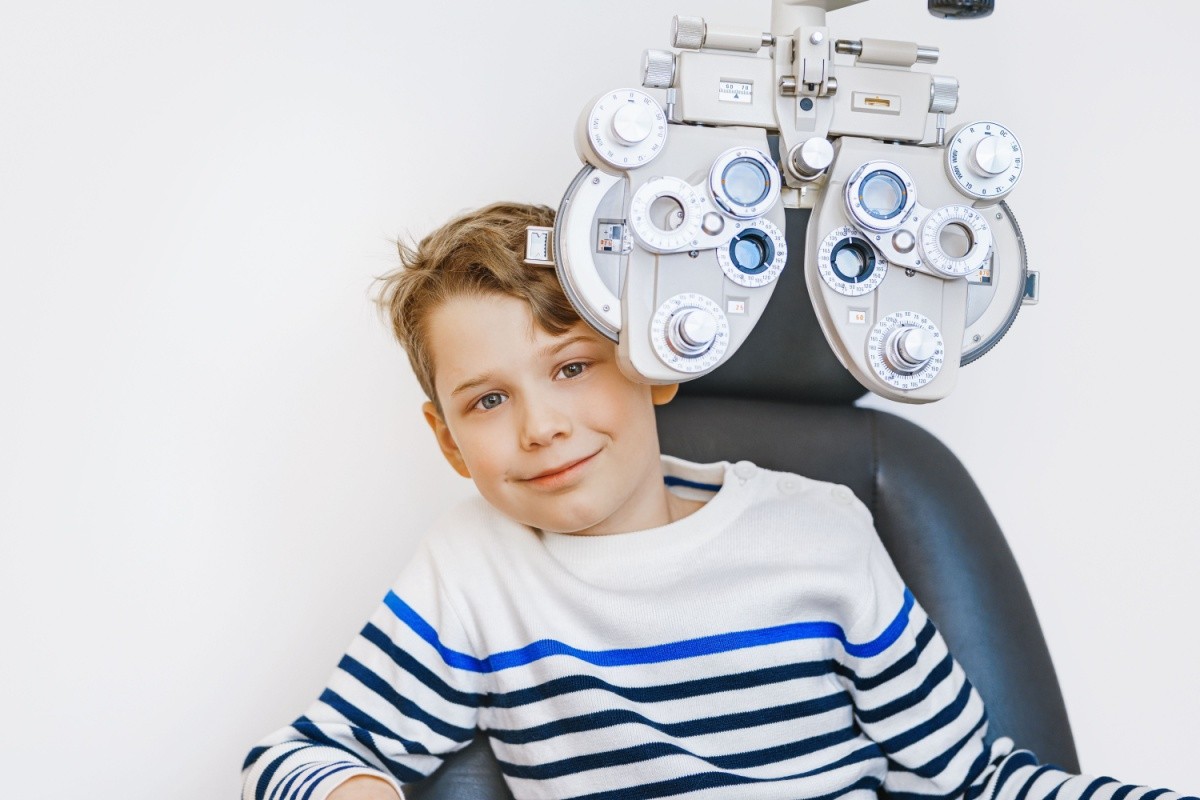 A young boy with brown hair sitting in an optometrist's chair, smiling, with a phoropter above his head.